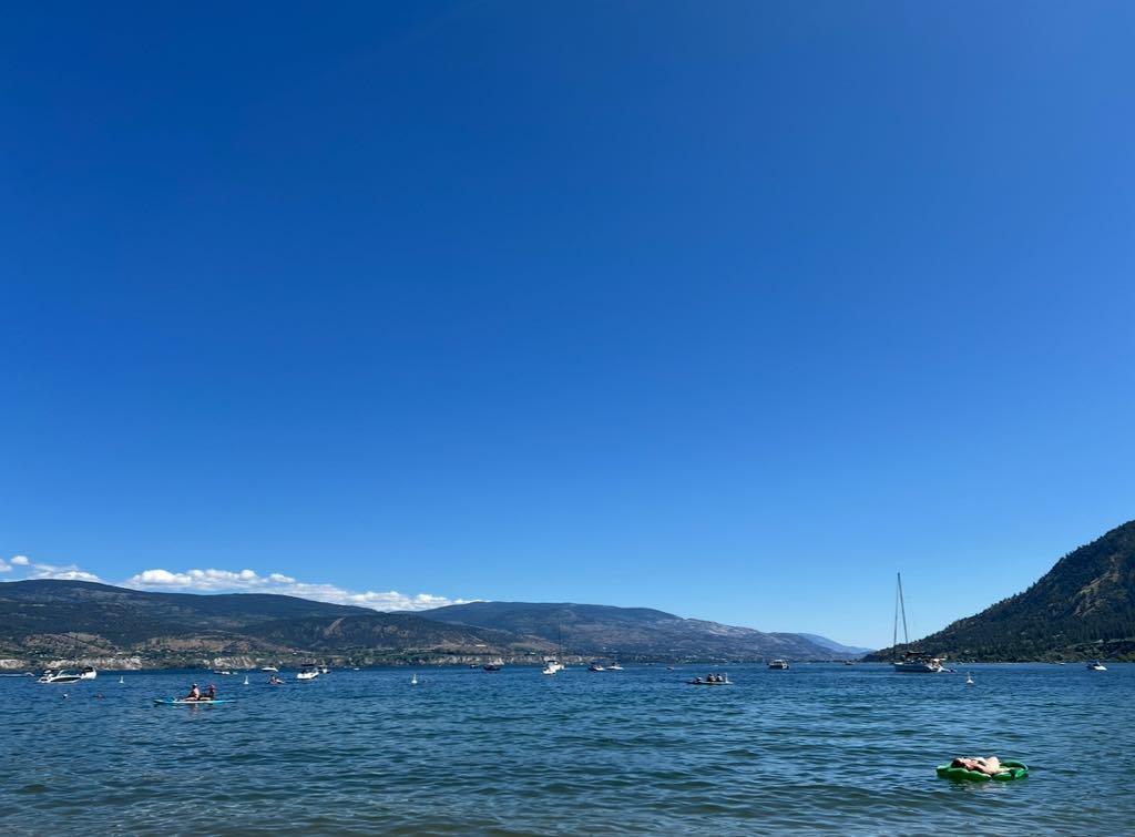 A peaceful summer day at the beach in Summerland, overlooking the clear blue waters of Okanagan Lake with mountains in the background.